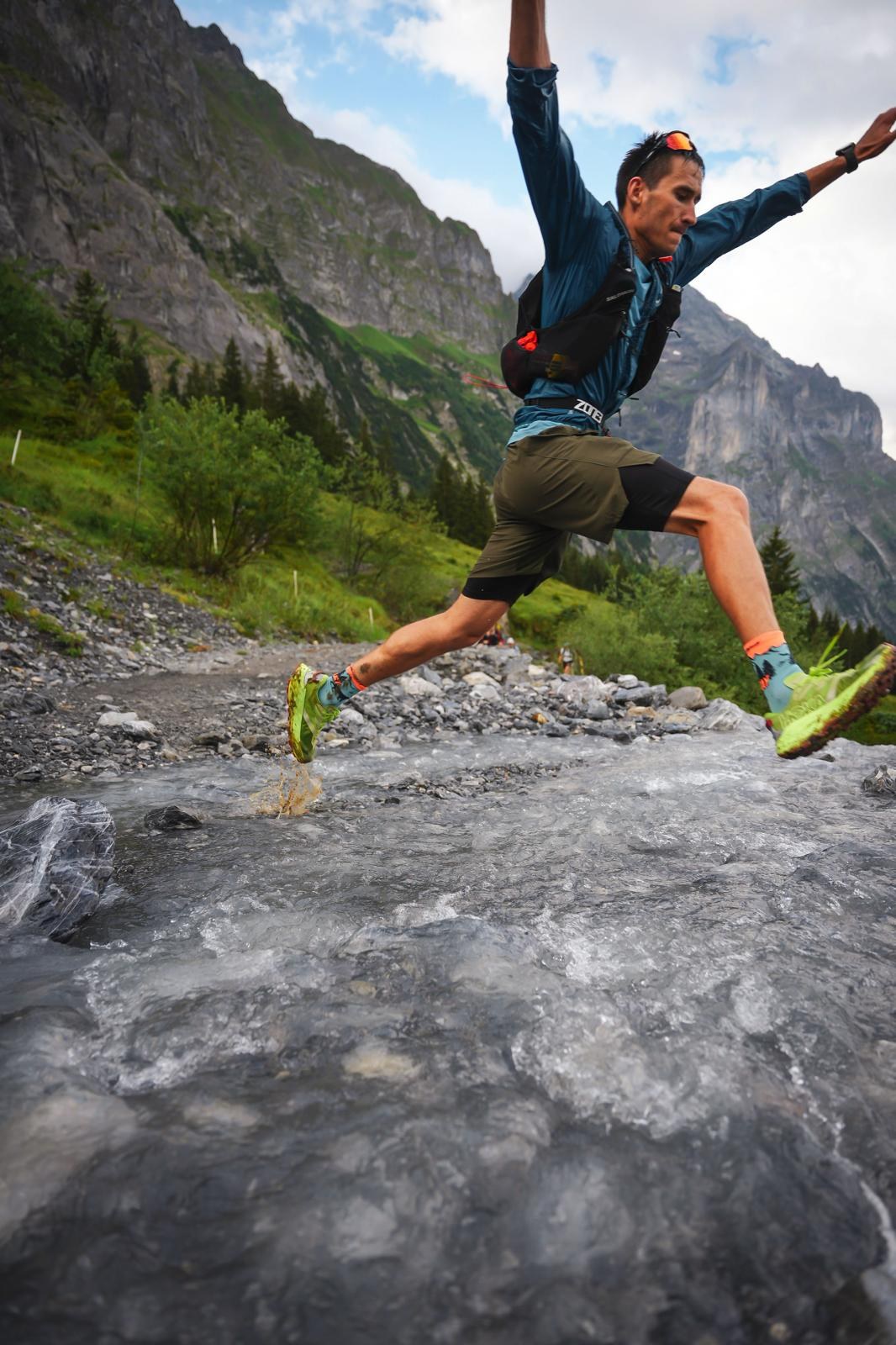Trail runner in a hydration vest leaping across a rocky mountain stream